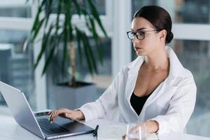 Caucasian middle-aged mixed-race businesswoman in glasses writing notes while sitting at desk in the office. People and wireless technology concepts. Working on laptop writing letter in paper notebook photo