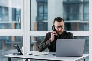 Smiling businessman holding smartphone sitting in office. Middle aged manager ceo using cell phone mobile apps and laptop. Digital technology applications and solutions for business development. photo