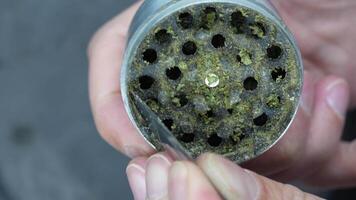 Close-up of a person cleaning a metal herb grinder with a tool, removing residue from clogged grinding holes, against a blurred dark background video