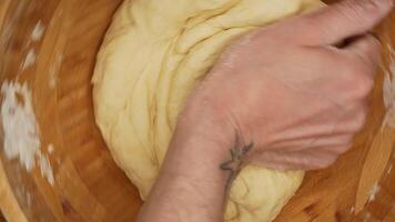 Close-up of tattooed male hands kneading dough in a wooden bowl, fingers covered in flour, veins visible, rustic wooden surface, warm and soft kitchen lighting creating a cozy and homely baking video