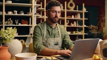 Serious Caucasian skilled man guy small business owner businessman crock potter ceramist artisan male working typing laptop networking computer online shopping ordering material inside workshop studio video