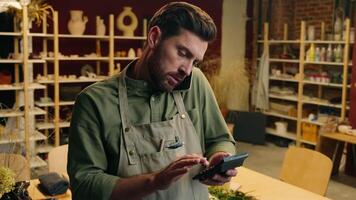 Busy Caucasian man male guy small business owner businessman potter craftsman using calculator counting finance calculate price money talking mobile smartphone phone call client order inside workshop video