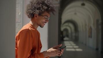 Happy smiling positive African American man holding mobile phone indoors side view joyful ethnic guy male student user gadget addict using smartphone in university college corridor social media chat video