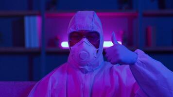 A man dressed in a protective suit sits focused while working online, surrounded by modern decor and vibrant neon lights, reflecting the challenges faced during the pandemic. video
