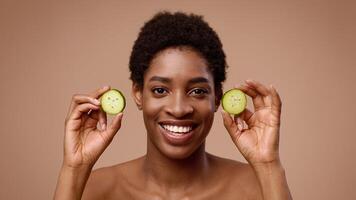 A cheerful young African American woman holds cucumber slices by her face while smiling. She embraces a moment of relaxation and self-care in a softly lit, isolated background. video