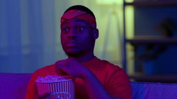 A young African American man relaxes in a cozy living room surrounded by modern decor. He enjoys a movie while snacking on popcorn in a vibrant atmosphere. video