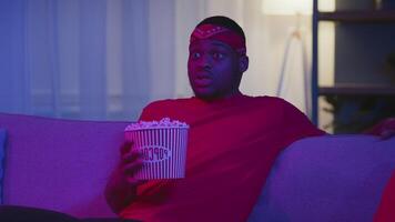 A young African American man is engaged in work at home during the night. He wears a vibrant red shirt and a matching headband, snacking on popcorn in a stylish room filled with neon lights. video