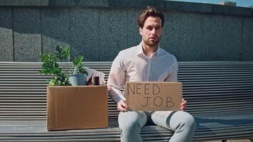 A young Caucasian man sits on a bench outside, holding a cardboard sign that reads Need Job. Next to him are boxes containing plants and office supplies, reflecting his struggles. video