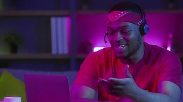 An African American man is working on a laptop in his modern home, illuminated by colorful neon lights. He appears engaged and is communicating online, showcasing the remote work lifestyle. video