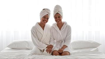 Two African American sisters sit together on a bed, wearing matching bathrobes with towels wrapped around their heads. They display warmth and connection in their family bond. video
