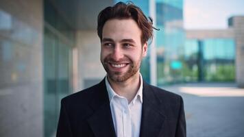 A millennial man stands outside a sleek corporate building, wearing a suit and smiling. His expression conveys optimism amidst challenges in his career as an entrepreneur and manager. video