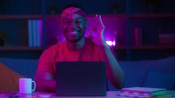A cheerful African American man sits at a desk in his home office, working on a laptop. The space is illuminated with neon lights, creating a lively, futuristic atmosphere during nighttime. video