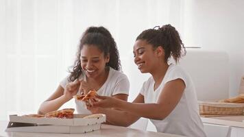 Two African American sisters are enjoying each other's company in a bright kitchen. They share laughter and conversation while savoring pizza, expressing their strong bond and love. video