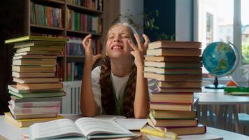 Angry frustrated irritated desperate Caucasian school little girl child student kid learner schoolgirl pupil sitting classroom desk between two piles books study problem hard difficult learning indoor photo