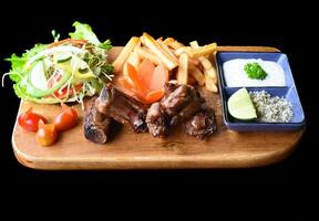Grilled smoked pork ribs with french fries and sauces on wooden board on a black background side view photo