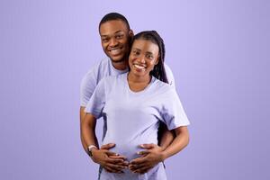 Portrait of smiling black guy hugging pregnant wife from back, cheerful couple standing over purple violet studio background, thinking about baby's name or gender. Empty speech bubble, free copy space photo