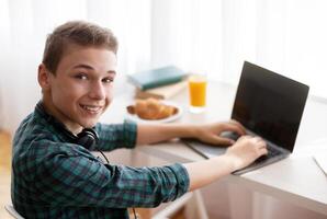 Cheerful schooler having break from studying at home, typing on laptop with empty screen, having snack photo