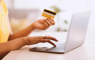 Making Financial Transaction. Closeup of unrecognizable black lady holding credit card and using laptop computer, typing on keyboard, sitting at desk, blurred background, selective focus on hand photo