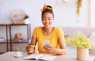 Online Order And Ecommerce Concept. Smiling African American Woman Using Smartphone And Holding Credit Card, Sitting At Desk With Open Notebook In Living Room At Home. Internet Shop Offers And Sales photo