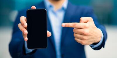 Modern technologies and business concept. Unrecognizable businessman in stylish suit pointing at smartphone with blank screen, recommending brand new mobile application for business, finance, closeup photo