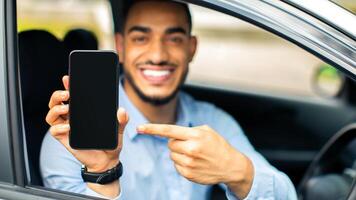 Smiling young arab businessman sitting in auto, pointing at smartphone with empty screen, showing newest mobile application for navigation, mockup. Car, automobile, driving and modern technologies photo