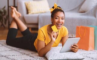 Online Shopping Concept. Happy Black Woman Using Digital Tablet, Holding And Showing Credit Card, Buying And Ordering Things In Internet Shop Lying On The Floor Carpet, Looking At Camera photo