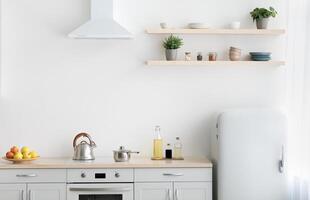 Stylish modern apartment with minimalist kitchen and Scandinavian interior. White furniture with stove, kettle and utensils. Fruits in plate, shelf with dishes and plants in pots, light wall photo