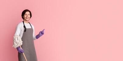 Cleaning Concept. Smiling man in apron holding mop and pointing at copy space on pink background, panorama photo