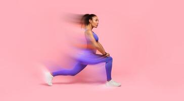 Young African American woman in activewear performing forward lunges on pink studio backdrop, displaying fitness routine, making stretching legs exercise looking aside, side view shot photo