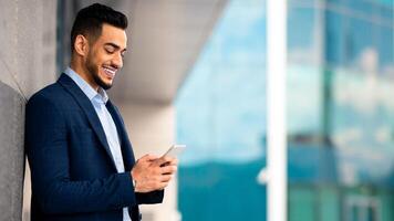 Handsome middle eastern businessman in suit standing by modern business center, using cellphone and smiling, sending emails while having break, panorama with copy space, side view photo