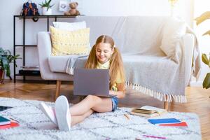 Modern Technology Concept. Full length portrait of happy teenage girl resting and sitting on the floor carpet, leaning on sofa, using her laptop, typing on keyboard, chatting or browsing web photo