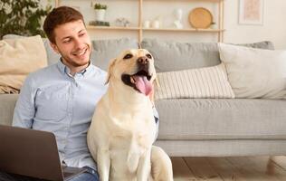Rest At Home Concept. Portrait of happy male owner of dog sitting on the floor in living room and patting his golden retiever. Guy holding computer on lap, spending time and relaxing at home photo