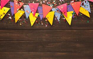 Border of colorful paper flags garland and confetti on rustic wood. Birthday party decoration background, top view, copy space. Mockup for greeting card photo