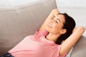 Rest Concept. Portrait of beautiful smiling lady lying on couch at home in living room. Happy lady relaxing on sofa, leaning back with closed eyes, enjoying weekend or break from work, free space photo