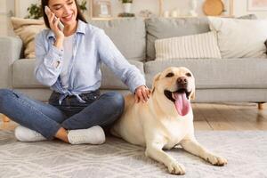 Everyday Life Concept. Casual girl talking on cell phone and caressing labrador, sitting on the floor in living room photo