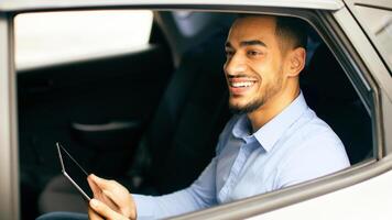 Cheerful middle-eastern businessman checking emails while going to airport by taxi, sitting on car back seat and using digital tablet, closeup photo, shot from outside, side view, copy space photo