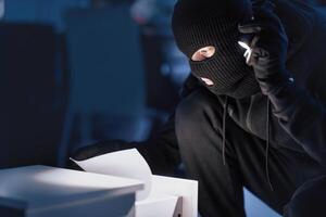 Closeup of disguised robber with flashlight searching for documents in office at night, copy space, blurred background photo