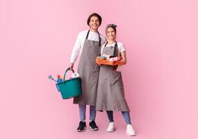 Professional Cleaners Team. Smiling Man And Woman In Aprons With Cleaning Tools In Hands Posing Over Pink Background With Copy Space photo