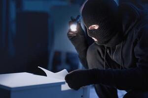 Industrial Espionage. Masked intruder holding and reading confidential documents using flashlight at night, copy space photo