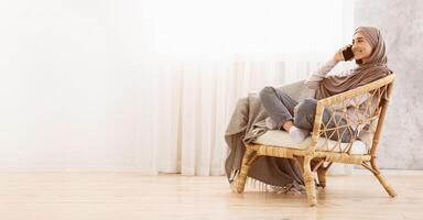 Muslim girl in hijab talking on cellphone while relaxing in cozy chair at home, sitting against window, side view with copy space photo