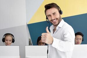 Happy male hotline operator smiling and showing thumb up gesture at modern open space office. Handsome tech support manager with headset expressing positivity and approval at call centre photo