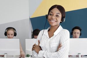 African American female tech support operator looking at camera and smiling at call centre. Portrait of helpdesk agent together with her colleagues working at modern office photo