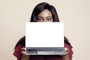 Young black lady holding notebook with empty screen, beige studio background, mockup, copy space. African american woman hiding behind computer with blank display for text or advertisement photo