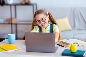 Distance Education. Smiling female pupil in eyeglasses sitting at desk, using laptop and typing on keyboard, doing homework, watching tutorial, lecture or webinar, studying online at home. E-learning photo