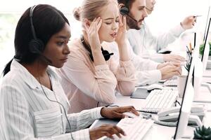 Technical support operator suffering from headache and her colleagues working with computers at office photo