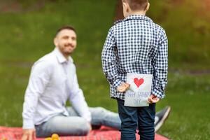 Father's Day surprise. Child hiding greeting card for dad behind his back on picnic in park, empty space photo
