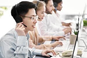 Asian helpdesk representative in headphones with his colleagues providing tech support services to customers, copy space. Team of call centre managers communicating with clients or selling online photo