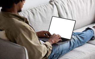 Black Man Using Laptop Computer With Blank White Screen While Sitting On Couch At Home, African American Guy Working Online Or Browsing Internet Website, Over Shoulder View, Mockup With Copy Space photo