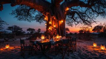 A beautifully arranged dinner table sits beneath a large baobab tree, illuminated by glowing lanterns, as the sun sets over the African savanna, creating a magical ambiance. photo
