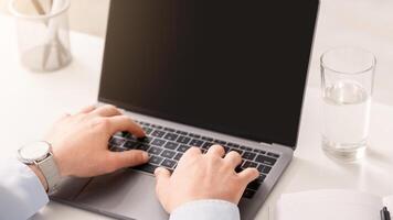 Man works at home. Hands of businessman typing on laptop with blank screen, on white table with glass of water photo